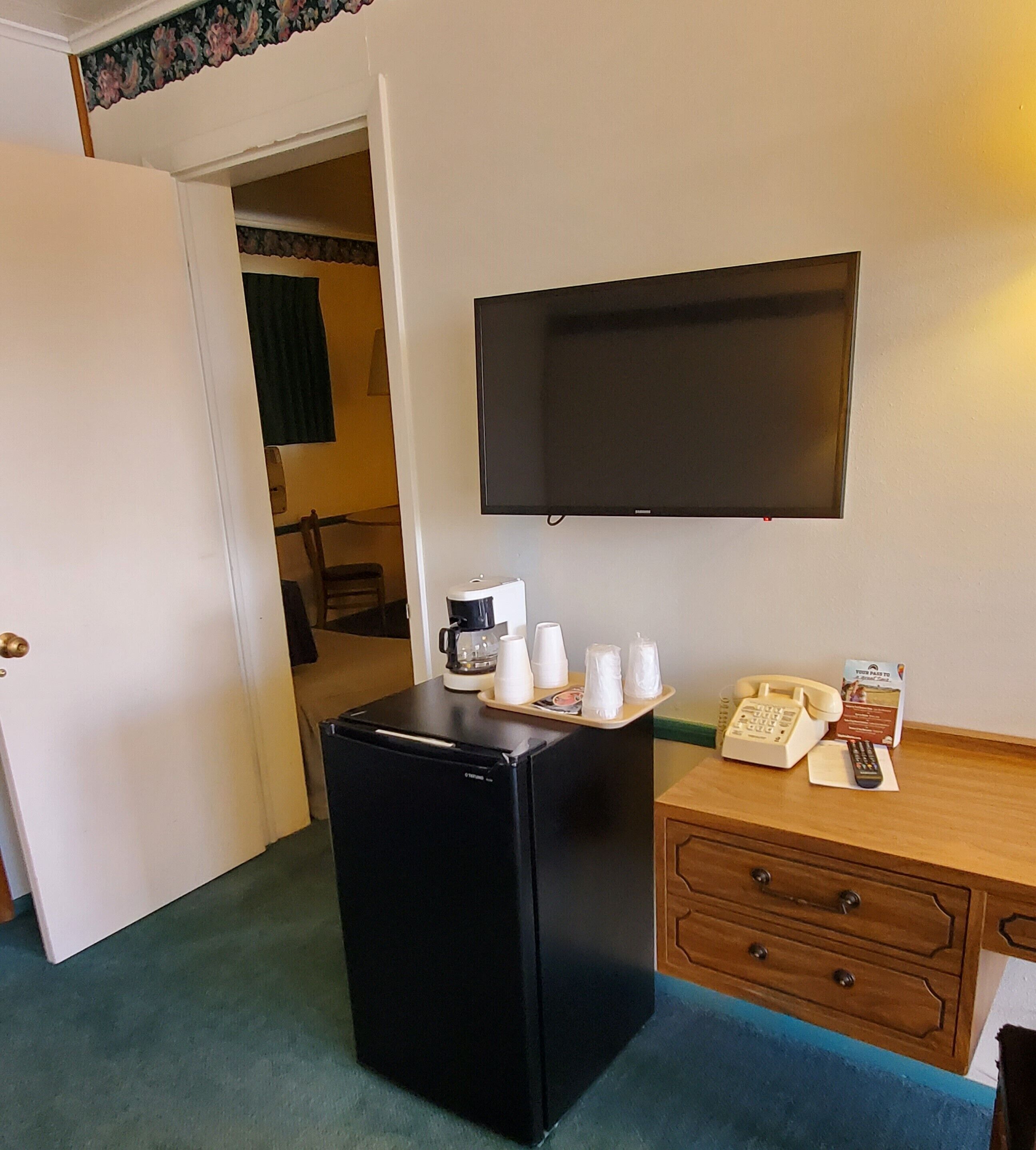 A compact hotel room with a black refrigerator, a coffee maker, a telephone, and a flat-screen TV mounted on the wall.