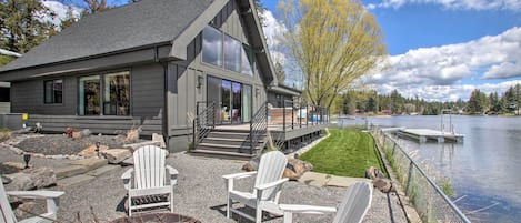 Una casa moderna con una terraza que da a un lago, con sillas Adirondack y un brasero.