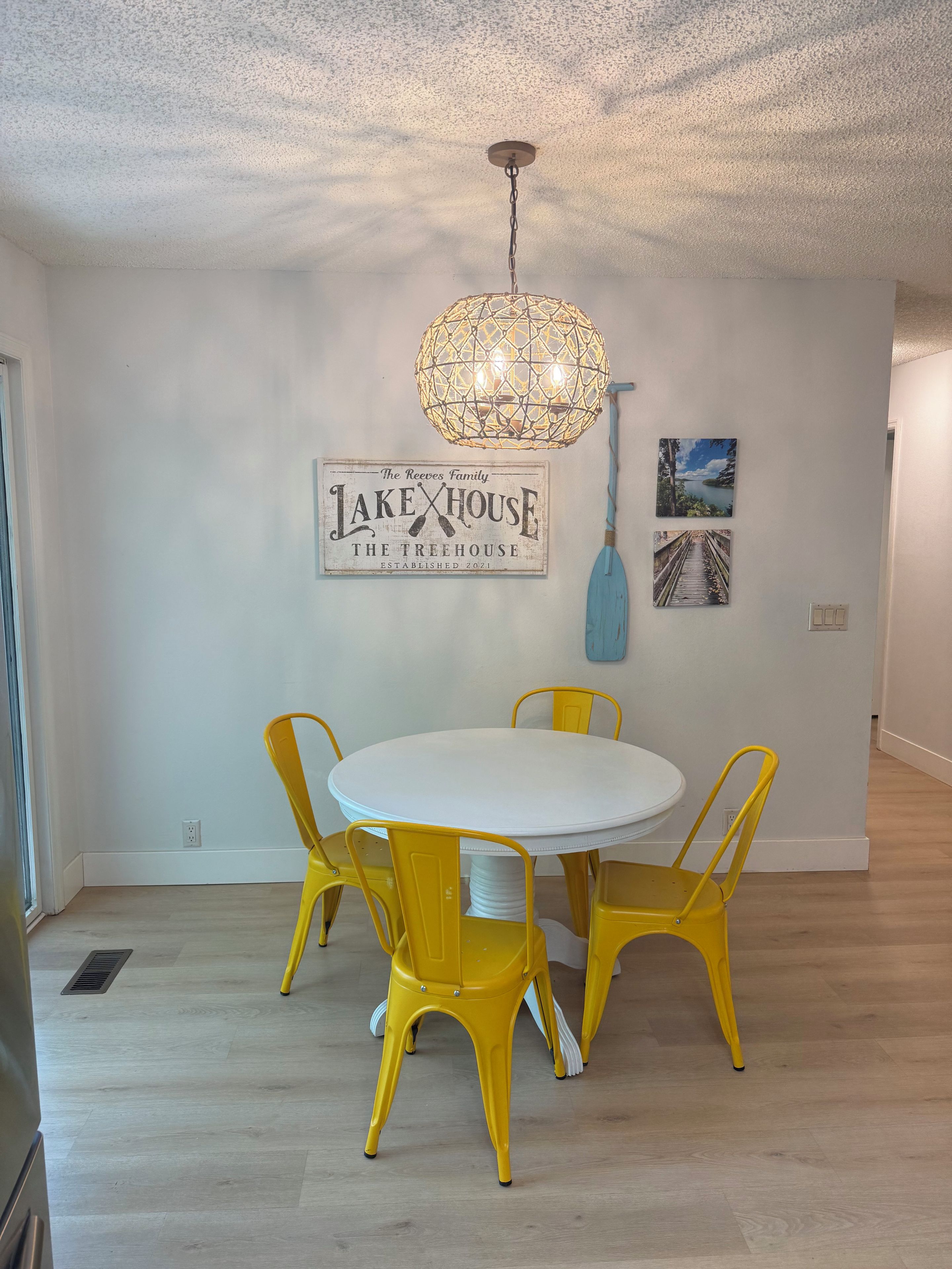 A dining area with a round table and yellow chairs, a decorative ceiling light, and wall art including a sign for 'The Reeves Family Lake House'.