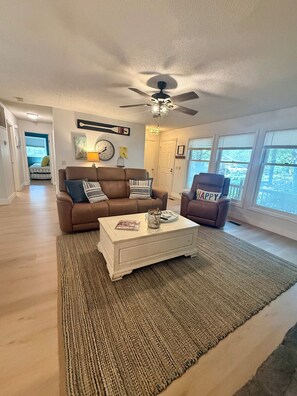 A spacious living room with a brown sofa, a white coffee table, and a ceiling fan. - The Treehouse - Cozy Cottage in the Woods (Hot Springs Village)