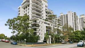 A multi-story residential building with balconies, surrounded by trees and parked cars.