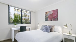 A hotel room with a bed, desk, chair, and a window with a view of buildings and plants.