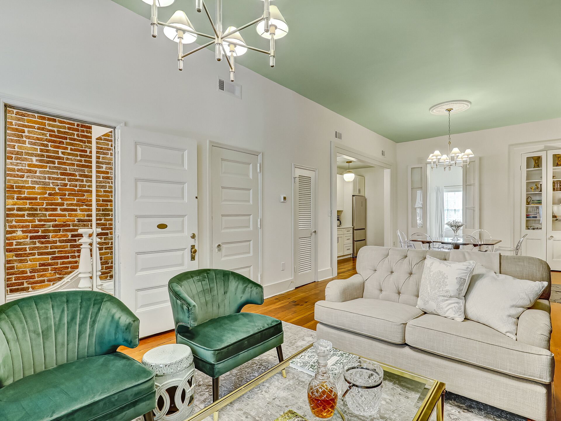 A spacious living room with a green sofa, a white sofa, and a coffee table with decorative items. There are two green armchairs, a brick wall, and a door with a brass handle.