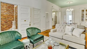 Amplia sala de estar con un sofá verde, un sofá blanco y una mesa de centro con objetos decorativos. También hay dos sillones verdes, una pared de ladrillo y una puerta con manija de latón.