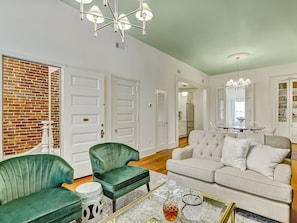 A spacious living room with a green sofa, a white sofa, and a coffee table with decorative items. There are two green armchairs, a brick wall, and a door with a brass handle. - Gorgeous Designer Apartment, Heated Pool Access (Savannah)