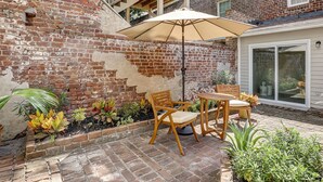 A patio with a brick wall, wooden chairs, an umbrella, and potted plants.