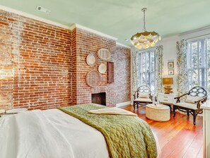 A room with a brick wall, a bed with a green quilt, two chairs, a small table, and a chandelier. - Gorgeous Designer Apartments, Heated Pool Access (Savannah)