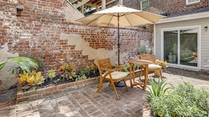 Un patio con pared de ladrillo, sillas de madera, una sombrilla y plantas en macetas.
