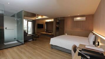 A modern hotel room with a large bed, a desk with a chair, a glass partitioned shower area, and a flat-screen TV mounted on the wall.