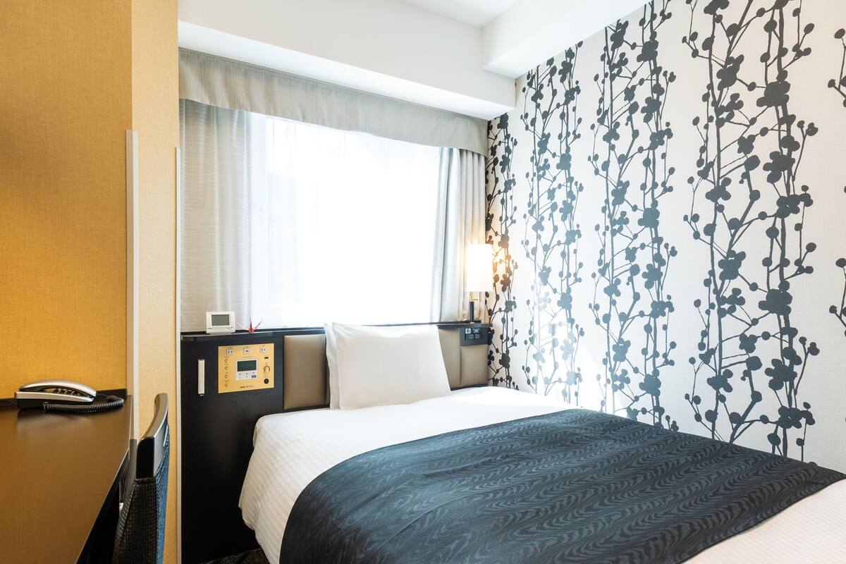 Une chambre d’hôtel avec un lit, une table de chevet avec un téléphone, un panneau de commande et une fenêtre avec des rideaux.