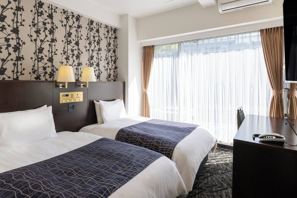 Une chambre d’hôtel avec deux lits, un bureau et une grande fenêtre avec des rideaux.