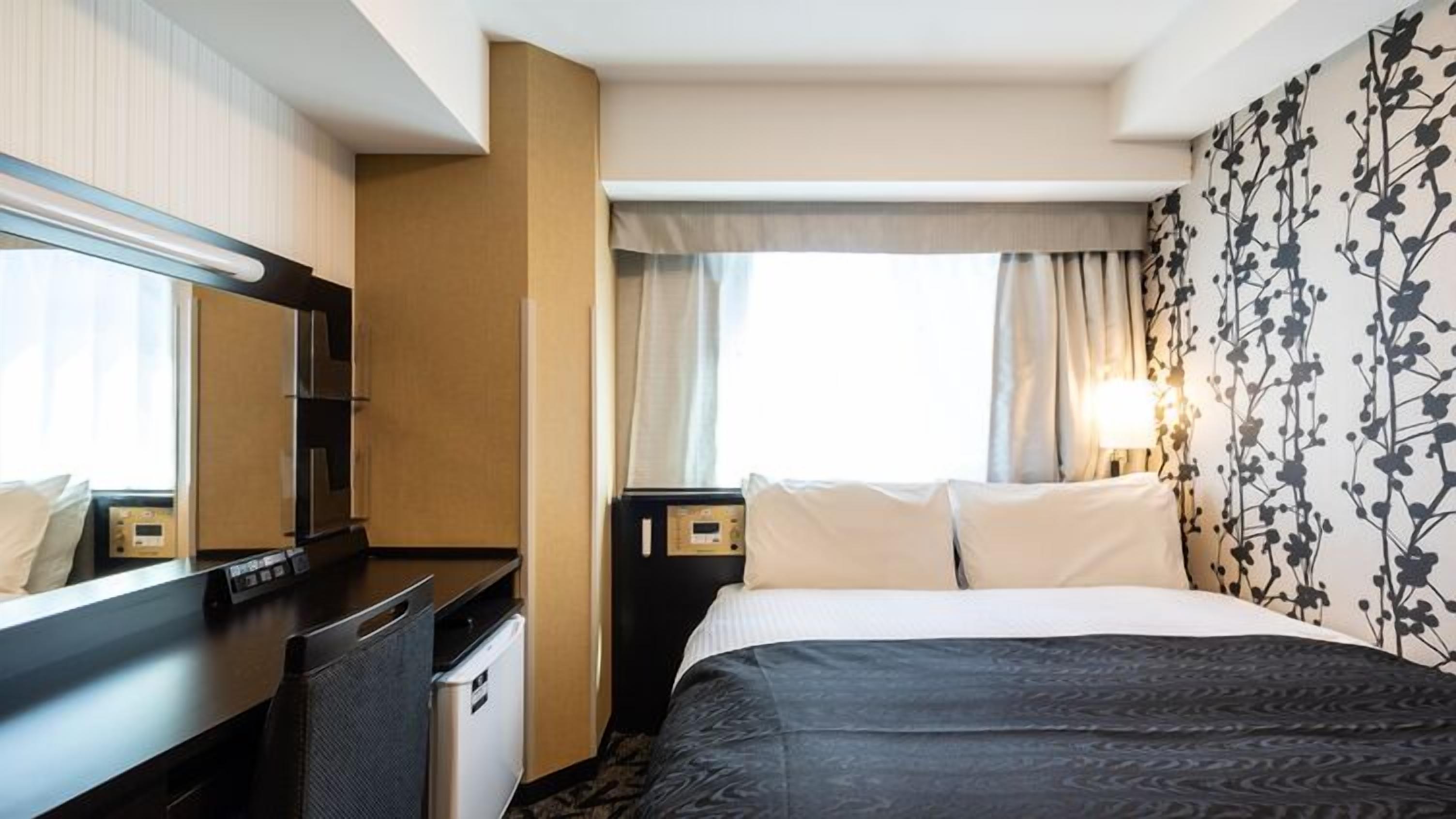 Una camera d'hotel con un letto, una scrivania, una sedia, un piccolo frigorifero, un comodino con una lampada e una finestra con tende.