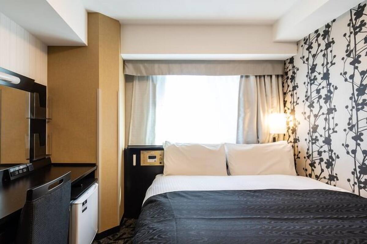 Une chambre d’hôtel comprenant un lit, un bureau, une chaise, un petit réfrigérateur, une table de chevet avec une lampe et une fenêtre avec des rideaux.
