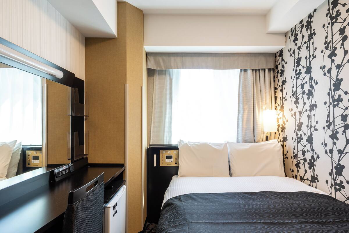 Une chambre d’hôtel avec un lit, un bureau, un miroir, une table de chevet et une fenêtre avec des rideaux.