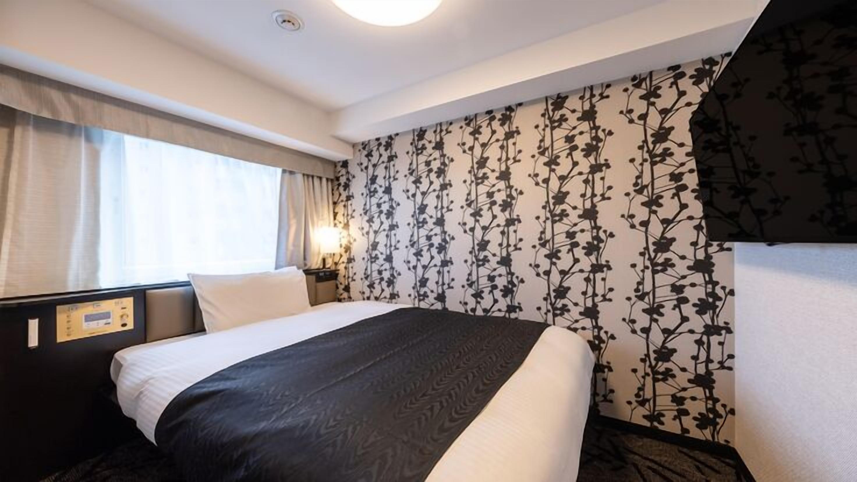 Camera d'albergo con un letto, un pannello di controllo, un comodino, una lampada e carta da parati con motivi floreali.