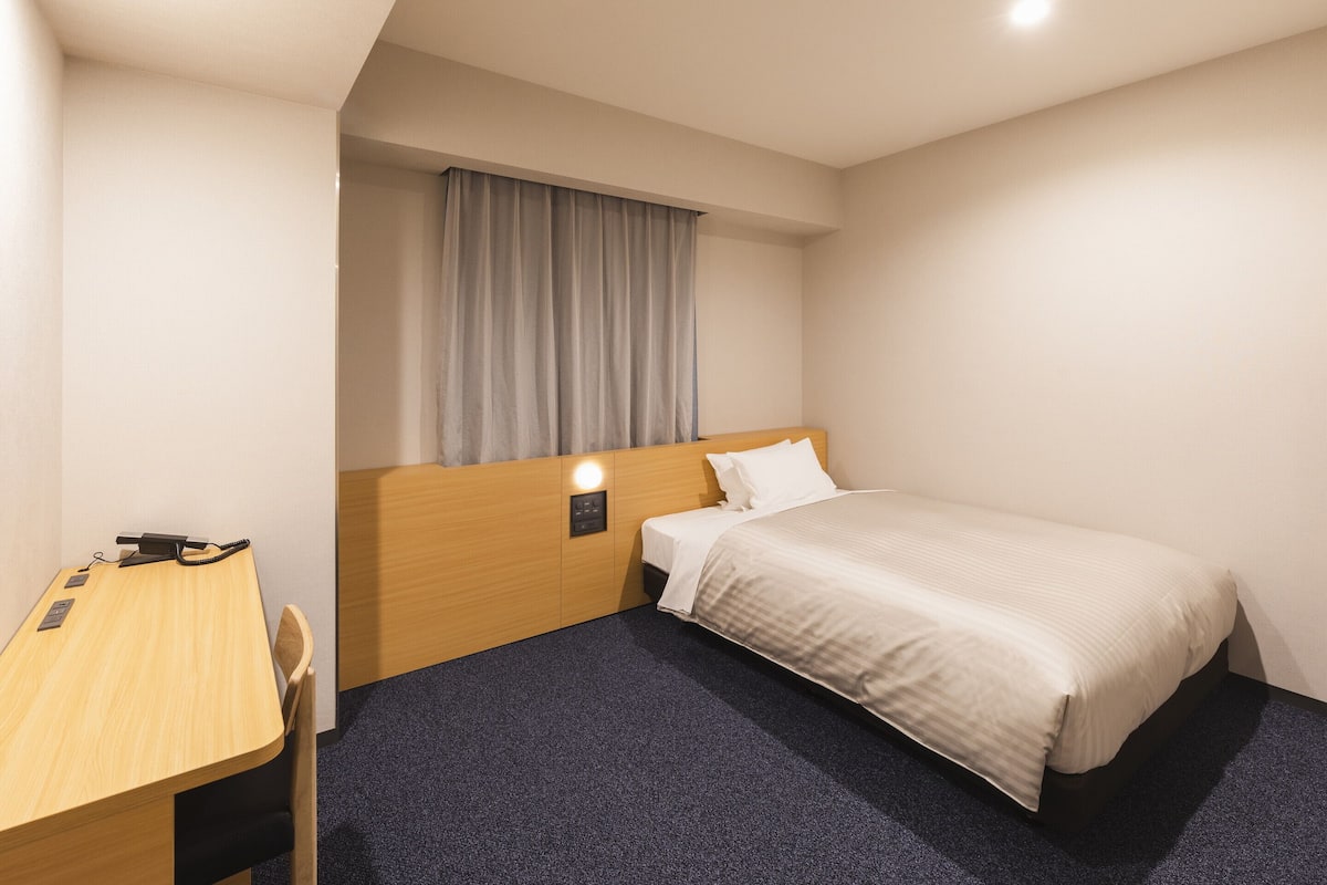 A hotel room with a single bed, a desk with a chair, a nightstand, and a window with curtains.