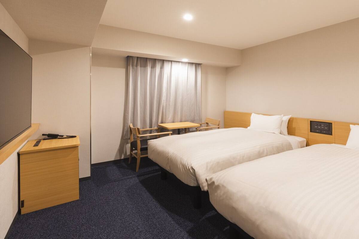 A hotel room with two beds, a desk, a chair, a small table, and a TV.