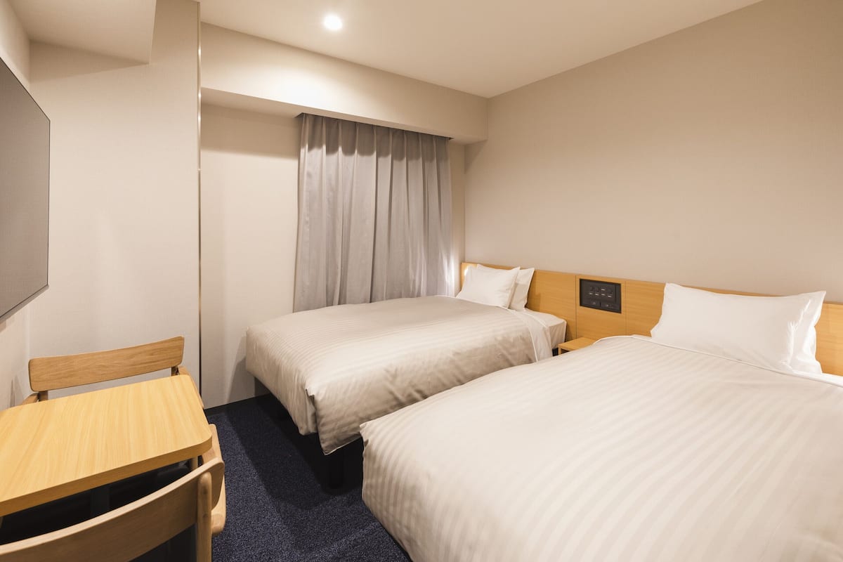 A hotel room with two beds, a wooden desk, a chair, and a large window with curtains.