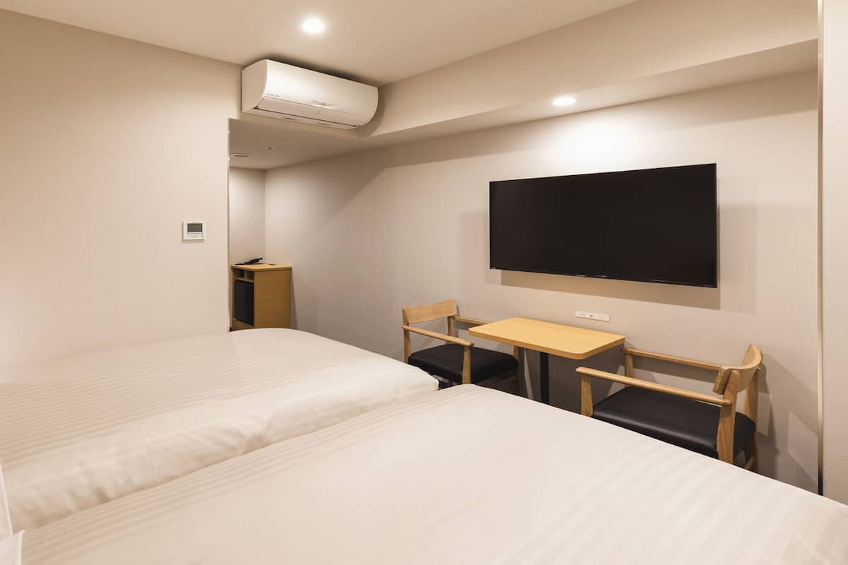 A hotel room with two beds, a desk, a chair, a flat-screen TV, and an air conditioning unit.