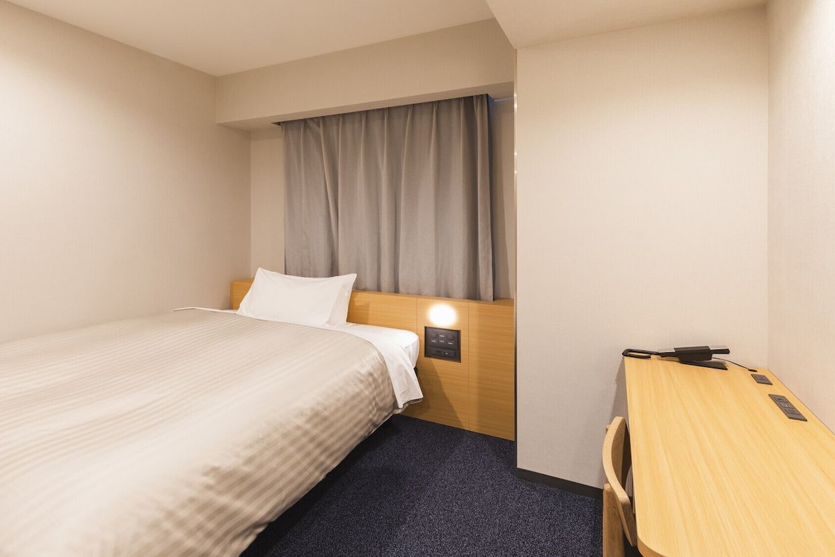 A hotel room with a bed, a desk, a nightstand, a wall-mounted lamp, and a window with curtains.