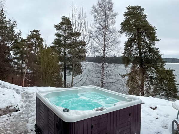 Ulkokylpyamme, jonka ulkopuoli on tehty puusta, ja joka sijaitsee lumipeitteisen maan ja puiden ympäröimänä, ja josta avautuu näkymä vesistöön.