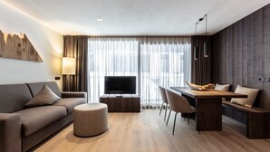 Ein modernes Wohnzimmer mit einer Couch, Hocker, Esstisch, Stühlen, einem Fernseher und einem großen Fenster mit Vorhängen.