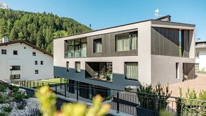 Ein modernes, zweistöckiges Gebäude mit großen Fenstern und einem Balkon, umgeben von einem angelegten Garten und einem bewaldeten Hang.
