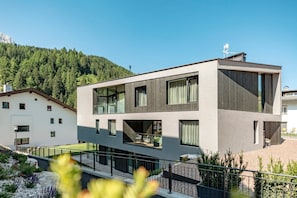 Un bâtiment moderne à deux étages, doté de grandes fenêtres et d’un balcon, entouré d’un jardin aménagé et d’une colline boisée.