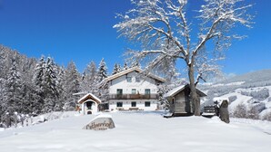 En snötäckt stuga med en trähusliknande byggnad, ett stort träd och en bergskedja i bakgrunden.