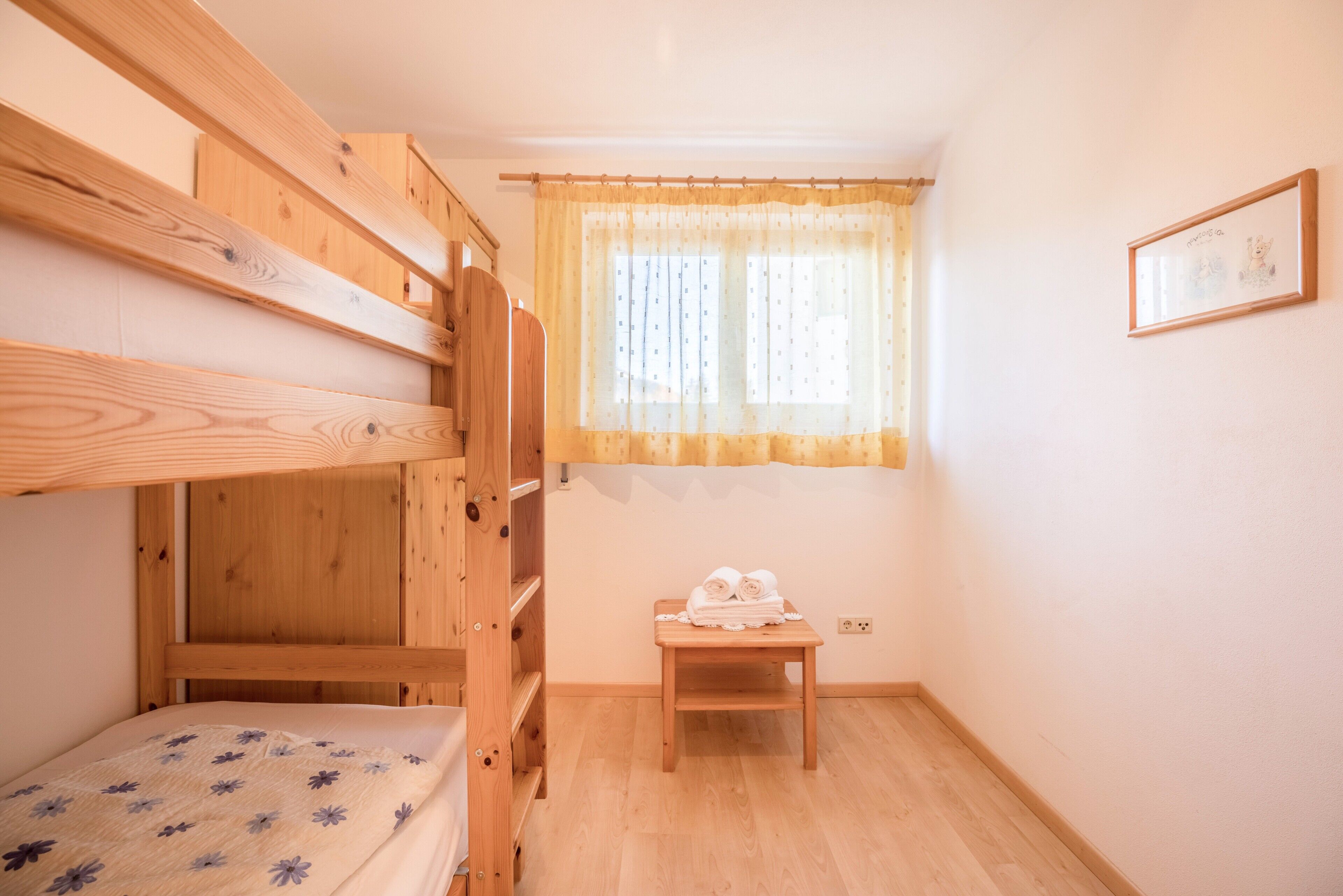 Ein Schlafraum mit Etagenbetten, einem hölzernen Kopfteil, einem kleinen Holztisch mit Handtüchern, einem Fenster mit Vorhang und einem gerahmten Bild an der Wand.