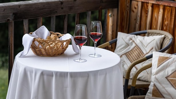 Una mesa redonda con mantel blanco, dos copas de vino, una cesta de mimbre y un paño blanco sobre una terraza de madera.