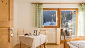 A room with a wooden door, a table with a white tablecloth, a bed, and a window with a view of a snowy landscape. - Lettnerhof – Family Suite Balcony 5. Surrounded by Nature, South-Facing Balcony, Mountain View, Wi-Fi. (Welsberg)