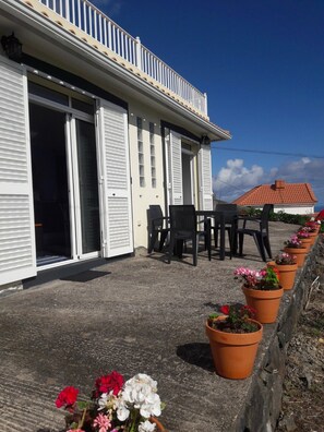 Un bâtiment à l’extérieur blanc, doté d’un balcon avec des chaises et des plantes en pot, et dont le toit est recouvert de tuiles rouges, visible au fond.