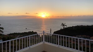 Vista do pôr do sol a partir de uma varanda com vista para a costa, casas e uma palmeira.