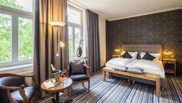 Ein Hotelzimmer mit einem großen Bett, einem Nachttisch, einem Sessel, einem kleinen Tisch mit einer Flasche und Gläsern sowie Blick auf Bäume durch das Fenster.