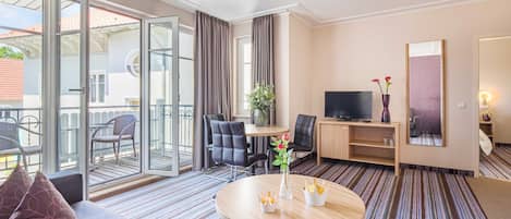 A hotel room with a sofa, a round table with a vase, a TV, a desk, and a chair.