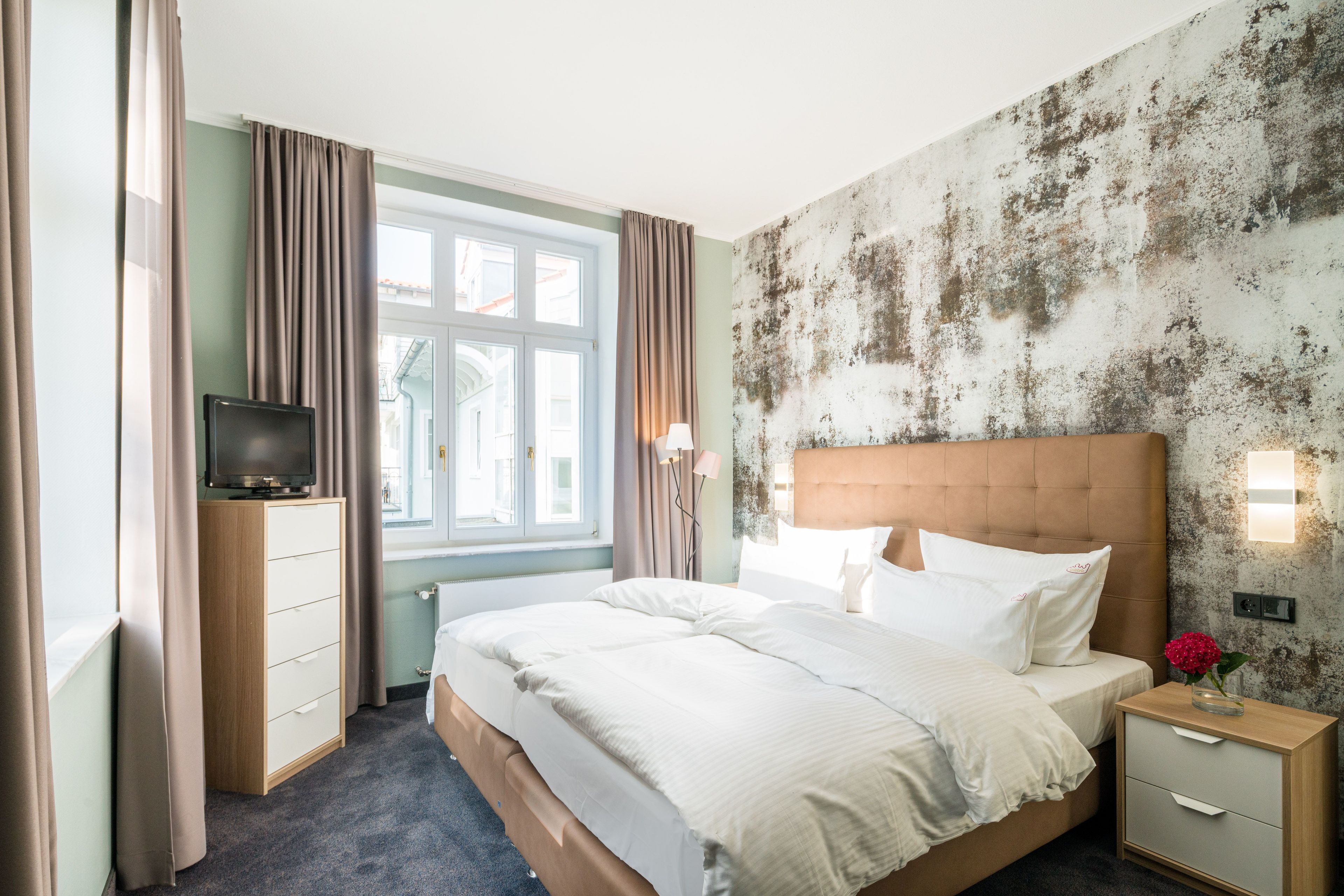 Ein Hotelzimmer mit Bett, Fernseher, Nachttisch, Lampe und einem Fenster mit Vorhängen.