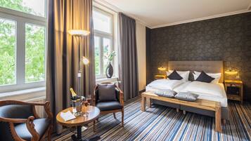 Ein Hotelzimmer mit einem großen Bett, einem Holzstuhl, einem kleinen Tisch mit Flasche und Gläsern sowie Blick auf Bäume durch das Fenster.