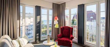 A room with a sofa, a red armchair, two small tables, and large windows with a view of buildings and a body of water.