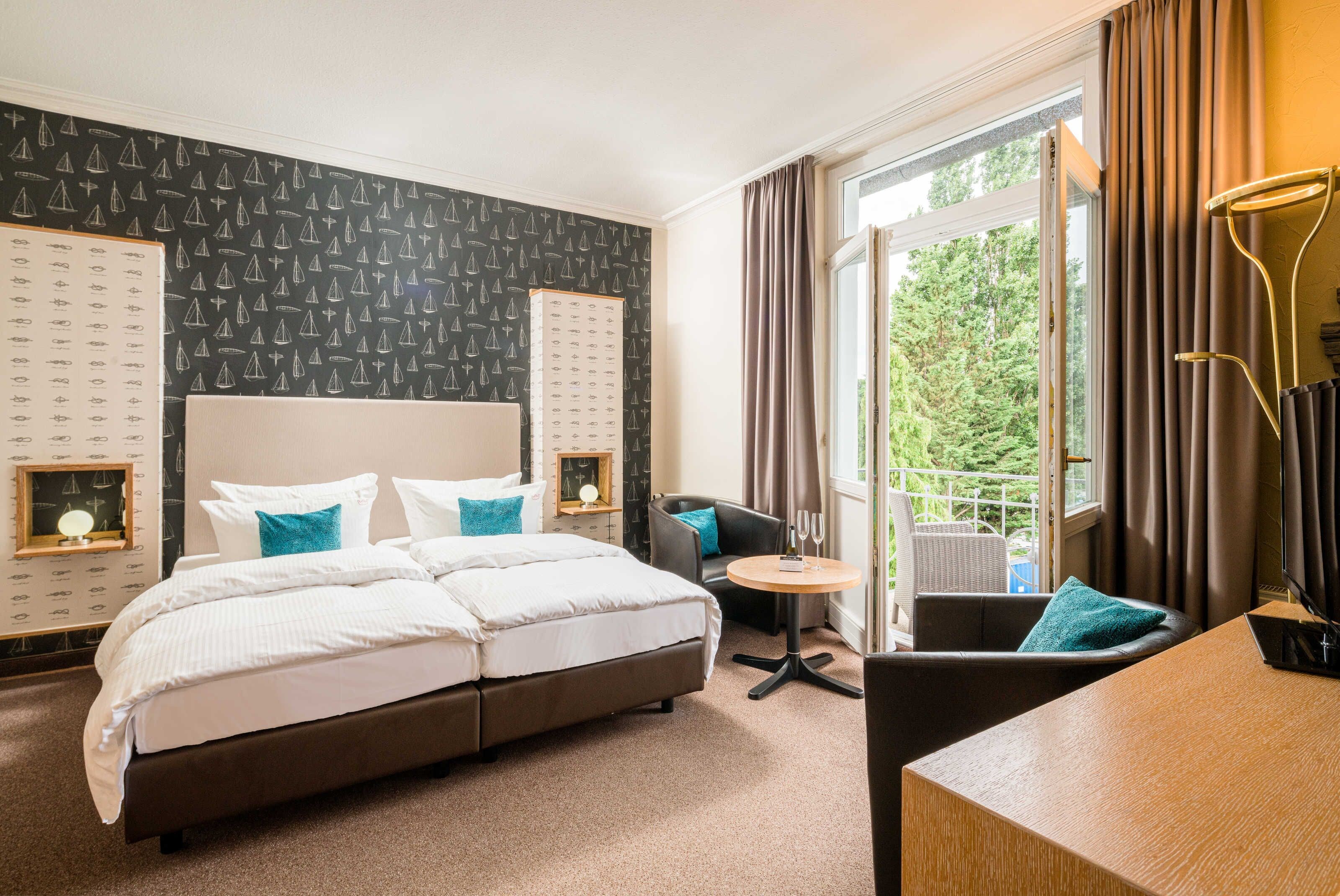 Ein Hotelzimmer mit einem großen Bett, einem Schreibtisch, einem Sessel, einem kleinen Tisch, einem Balkon mit Vorhängen und einer Wand mit Muster.