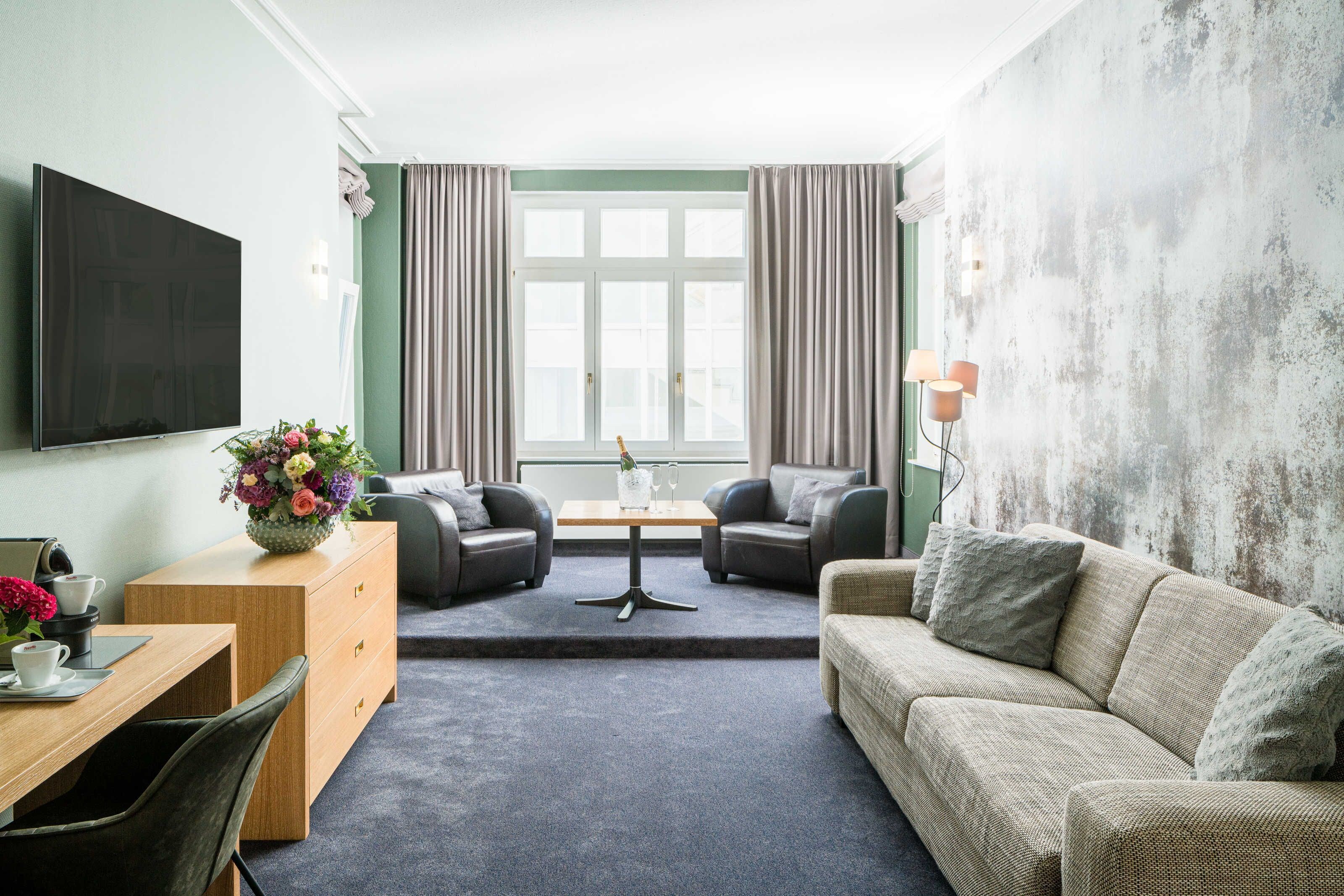 Une chambre d’hôtel moderne dotée d’un téléviseur à écran plat, d’un bureau en bois agrémenté d’un vase de fleurs, de deux canapés en cuir, d’une petite table et d’une grande fenêtre avec des rideaux.