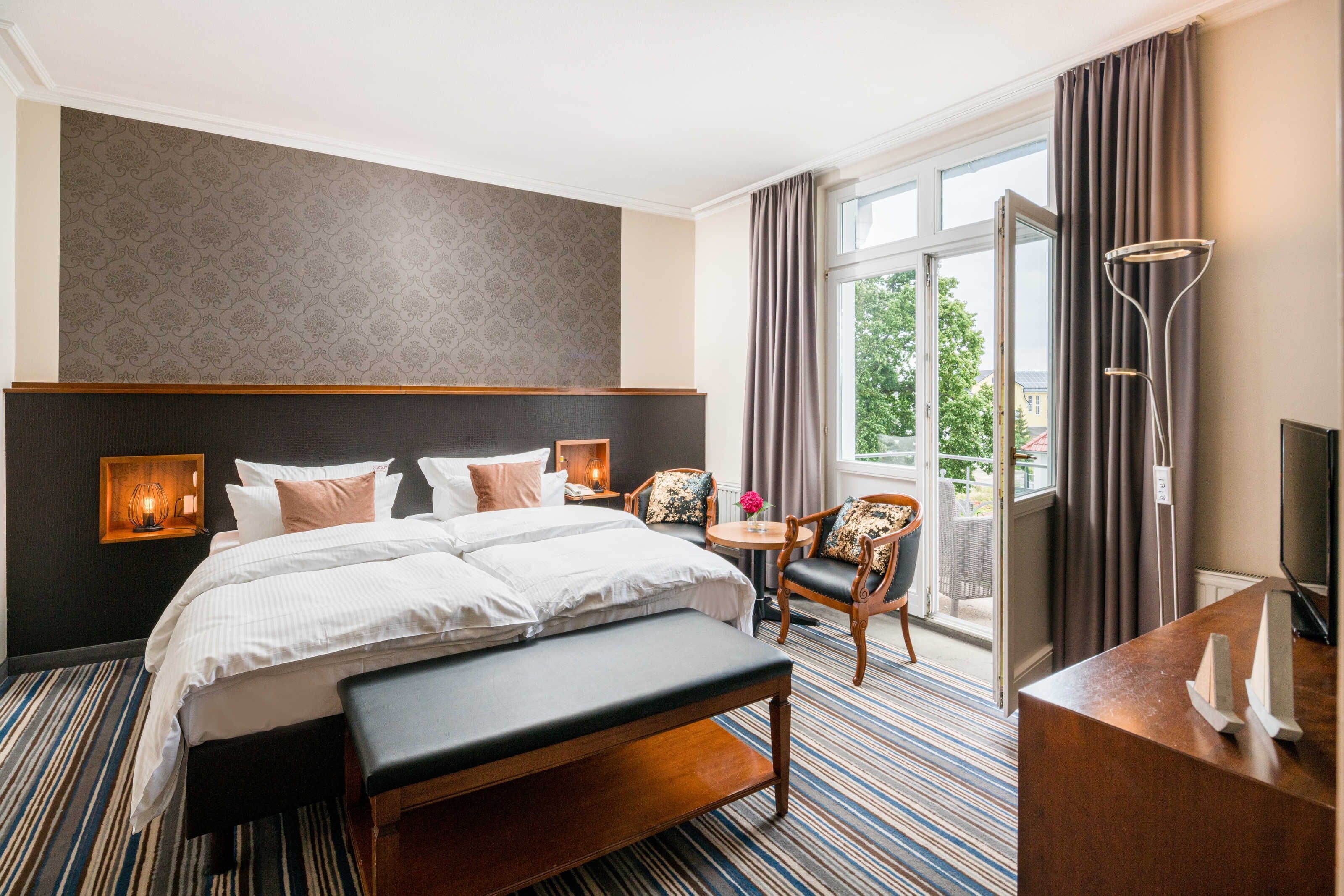 A hotel room with a large bed, a desk, a chair, a bench, a TV, and a balcony with curtains.