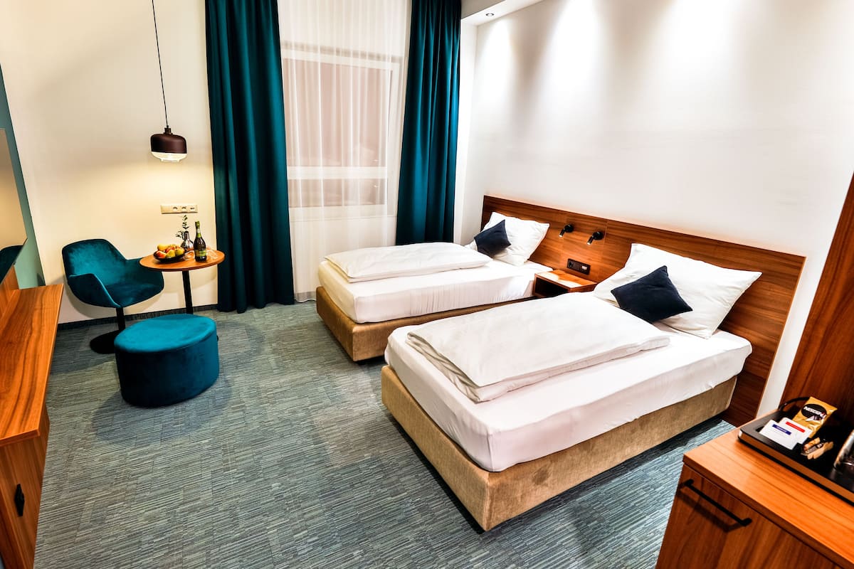 A hotel room with two beds, a desk, a chair, a small table with a bottle and fruit, and a window with curtains.