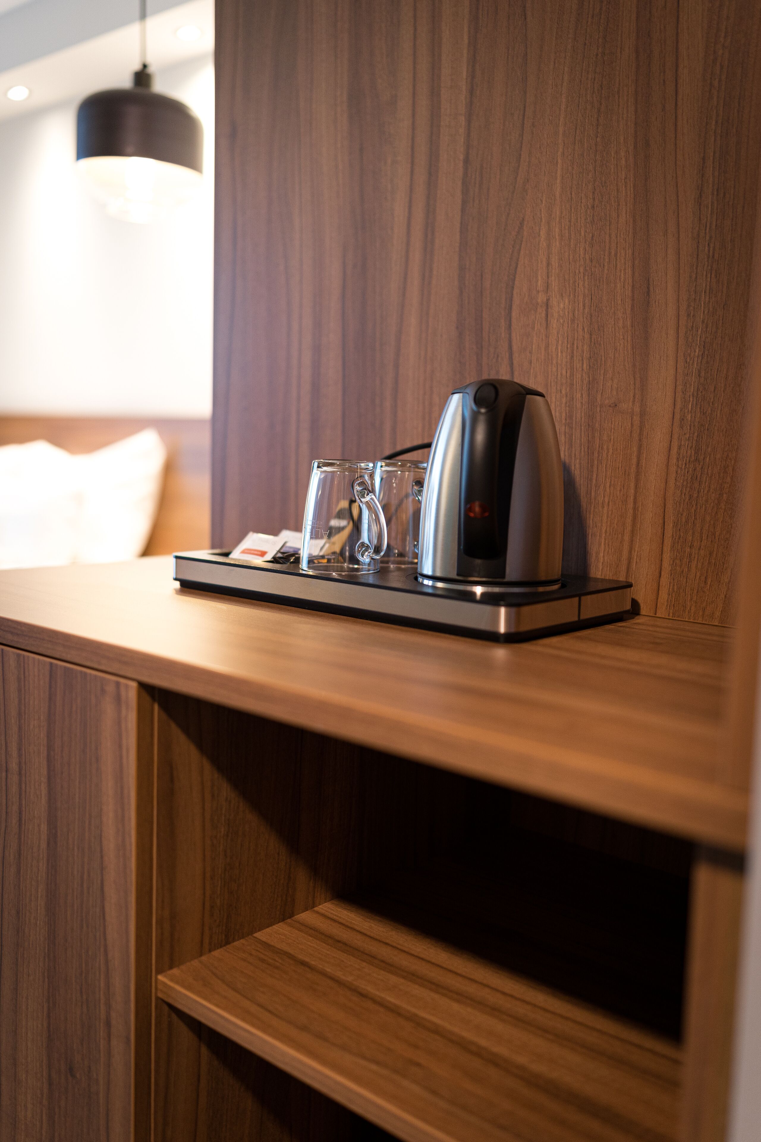 A coffee maker on a tray with a glass of water and a card, placed on a wooden surface in a room with a wooden wall and a hanging light fixture.