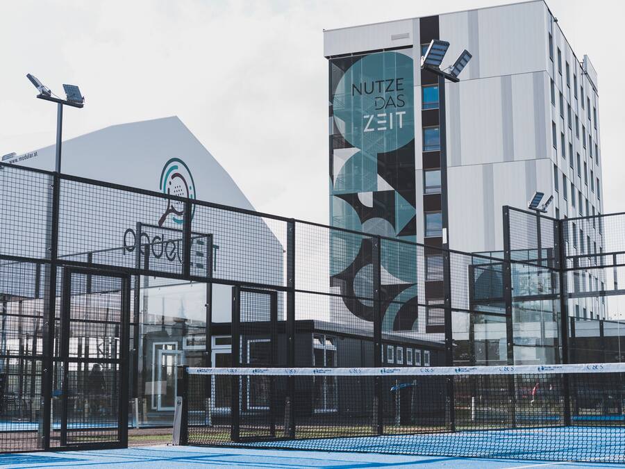 Instalación deportiva con cancha de tenis cubierta con red, un edificio grande con un logotipo y paneles solares en el techo.