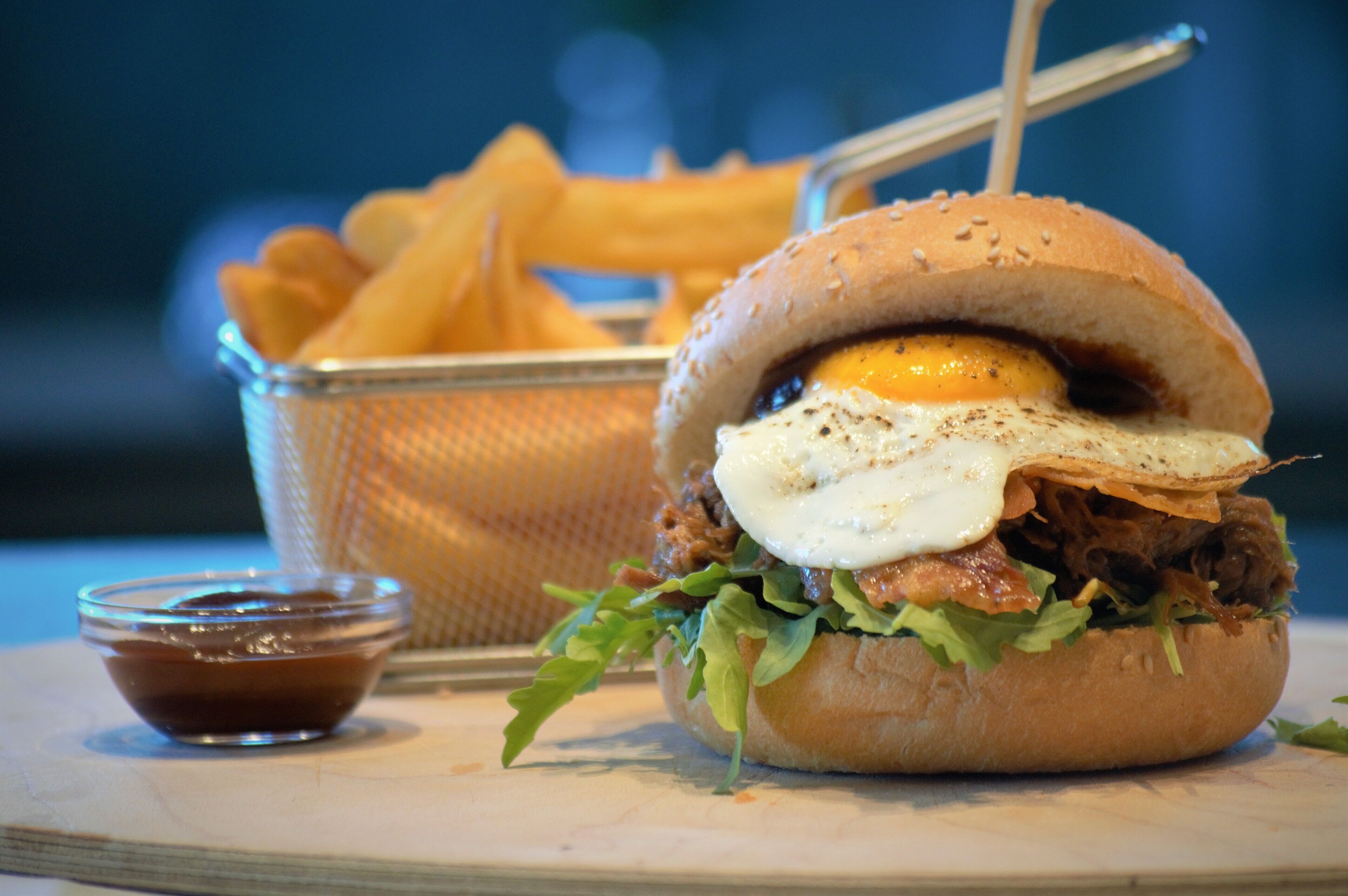 A sandwich with a fried egg, meat, and lettuce on a bun, accompanied by a basket of fries and a small bowl of sauce.