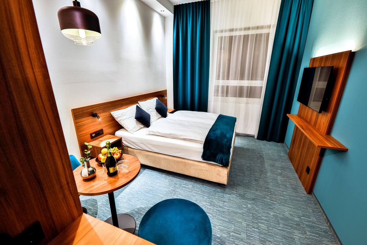 A hotel room with a bed, a round table with a bottle of champagne, a TV, and a blue chair.