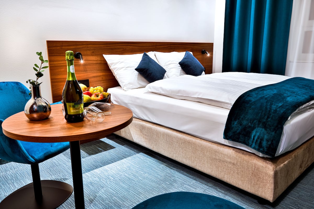 A hotel room with a bed, a round wooden table with a bottle and glasses, a blue chair, and a fruit bowl on the bedside table.