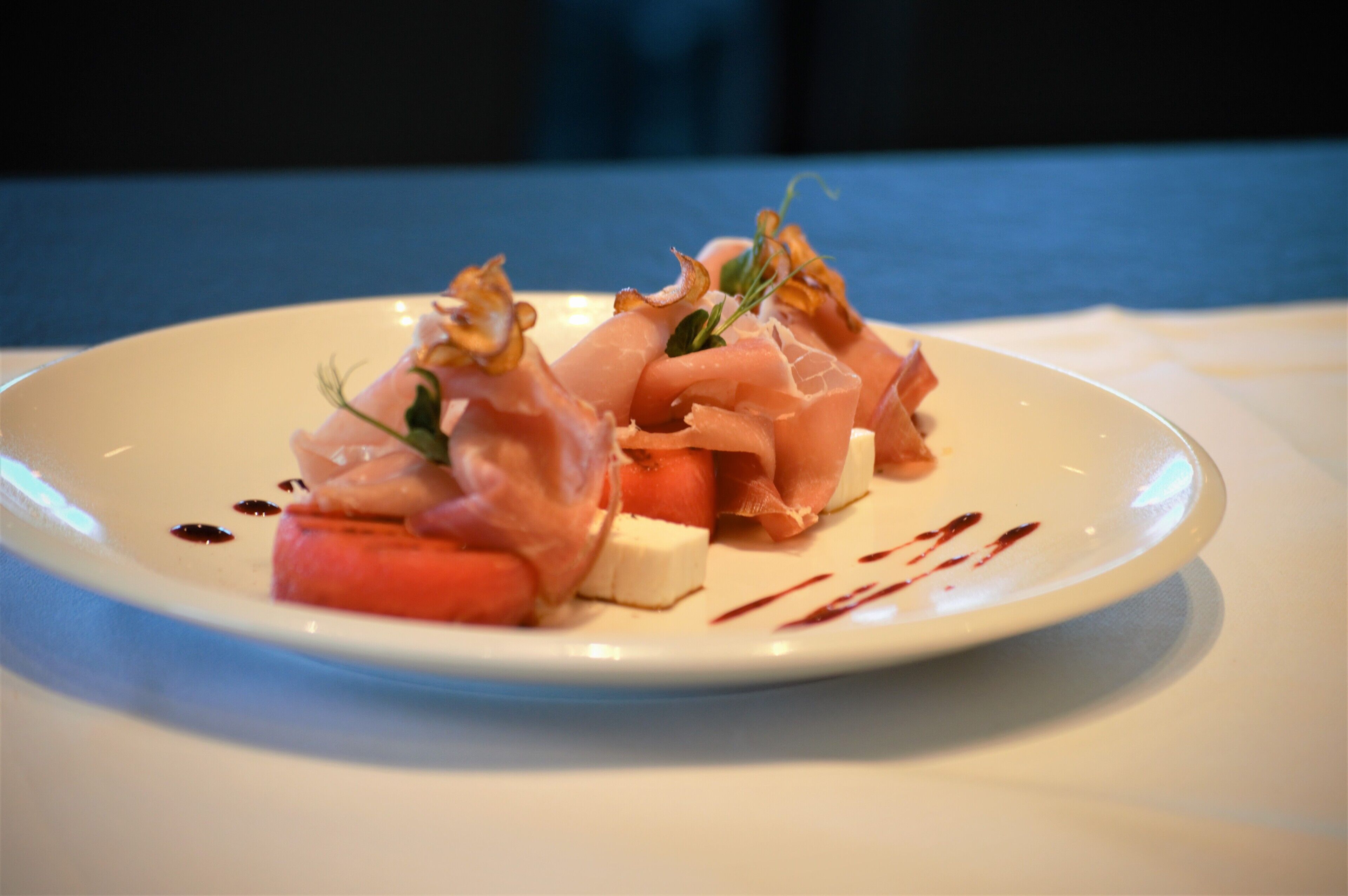 A white plate with a food arrangement including thinly sliced meat, a slice of tomato, and a dollop of cream cheese, garnished with a small green leaf.