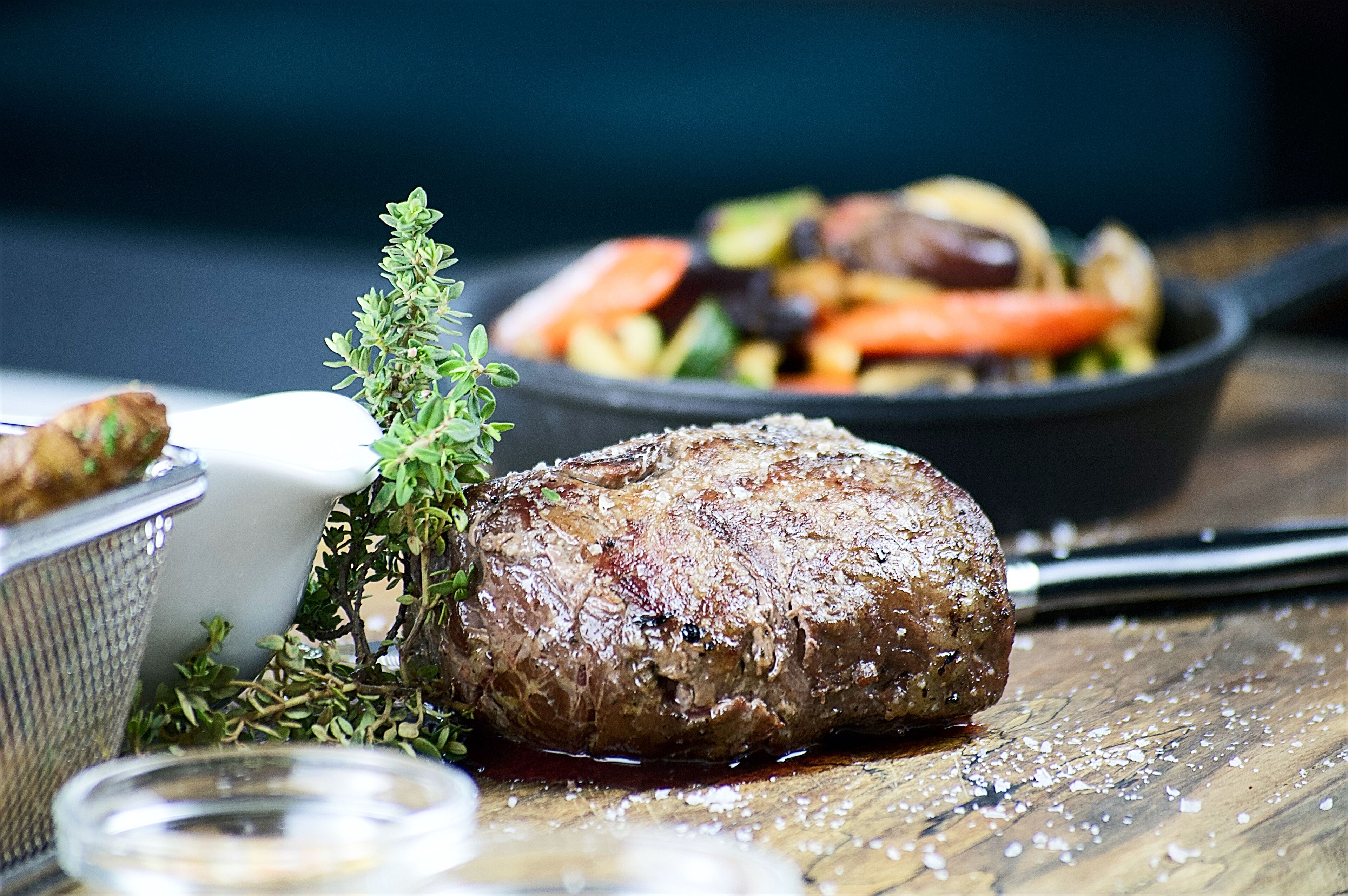 Ein Steak mit Thymianzweig auf einem Holztisch, begleitet von einer kleinen Schale mit Salz und Pfeffermühlen.